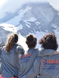 Trio of women wearing Reach Fitness hoodies sitting atop a mountain staring out over Mt. Hood.